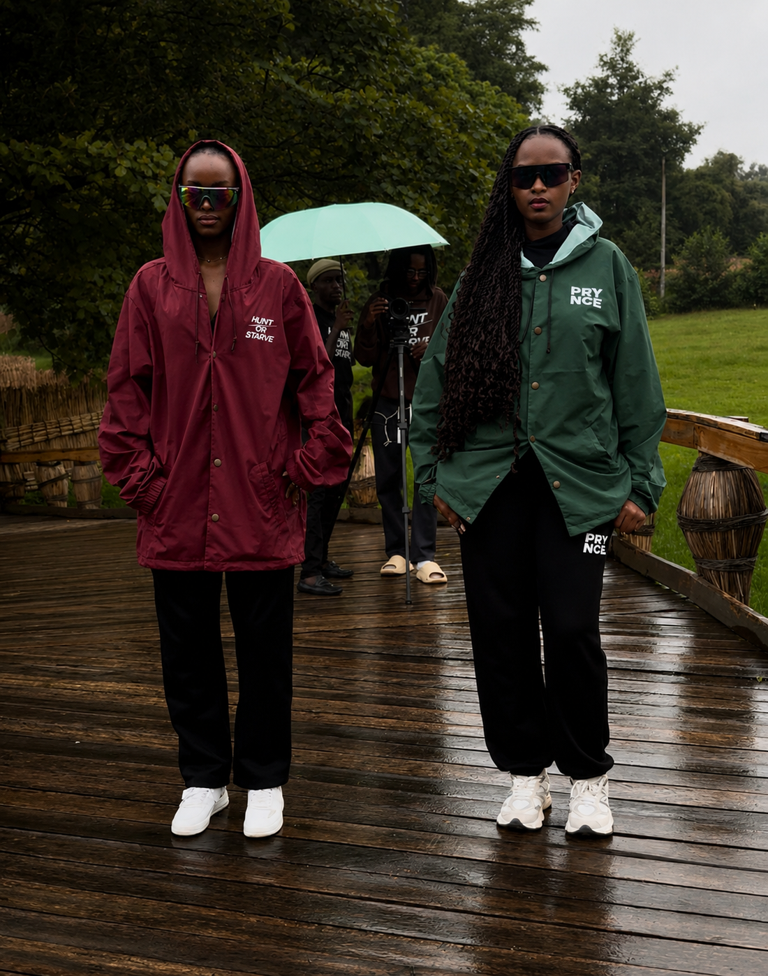 Two models wearing PRYNCE AFRICA Coach Jackets in burgundy and green on a rainy boardwalk in Kigali, Rwanda. Film crew visible in background.
